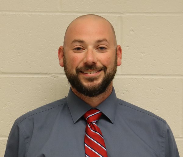 A smiling man with a beard and a red striped tie stands against a white brick wall.